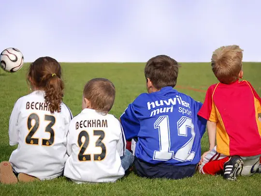 Four children in football jerseys sit on a grass field with a soccer ball