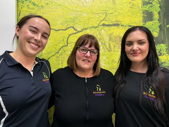 Three smiling Brindabella Podiatry team members in branded uniforms in front of a forest mural