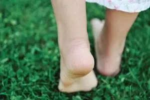 A child walks barefoot on grass, seen from behind at heel level