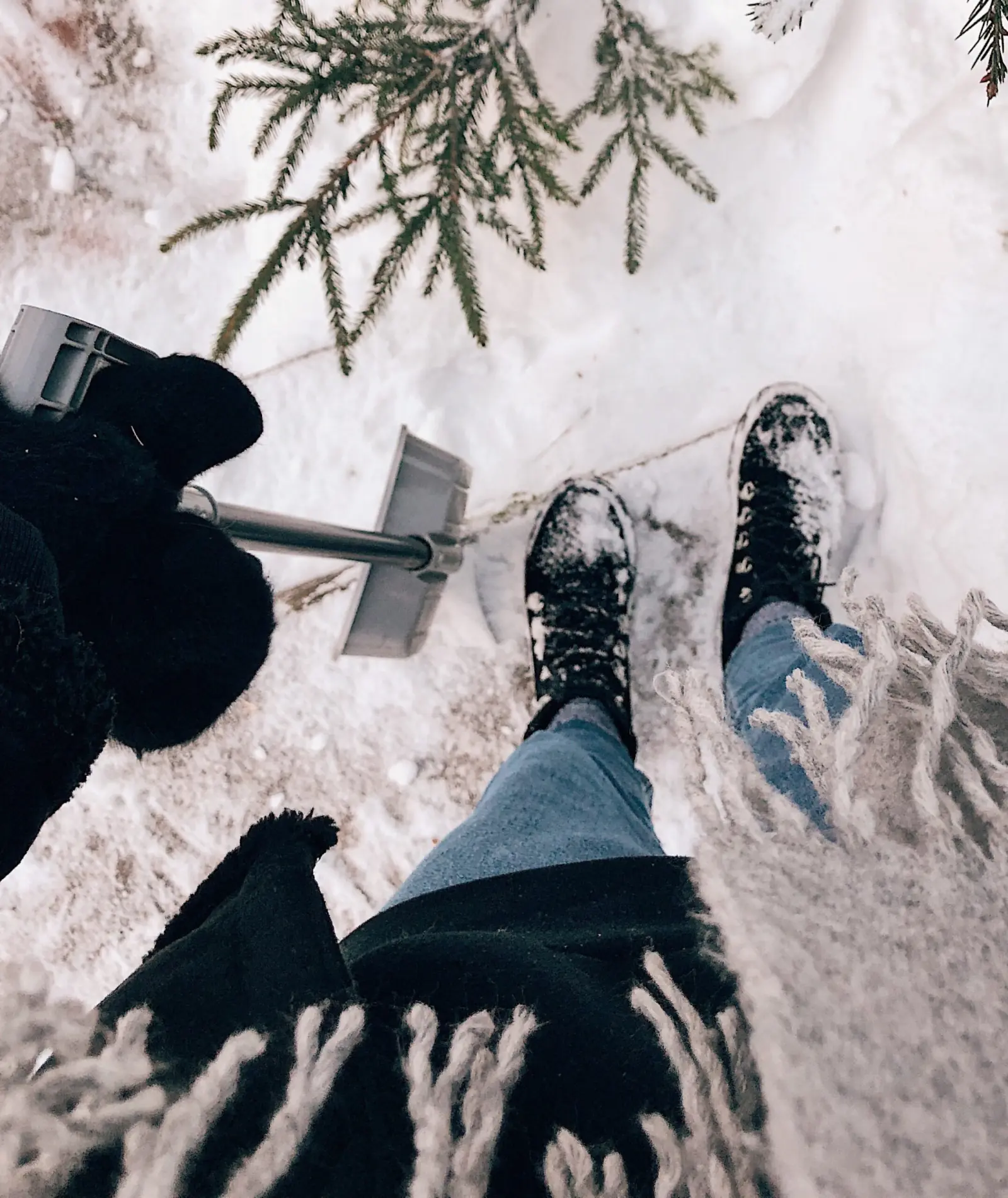 Looking down at snow-covered boots and jeans on a snowy path beside a snow shovel